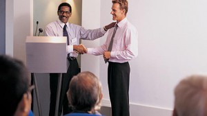 Businessman Shaking Another Businessman's Hand on a Podium During a Presentation in Front of an Applauding Audience