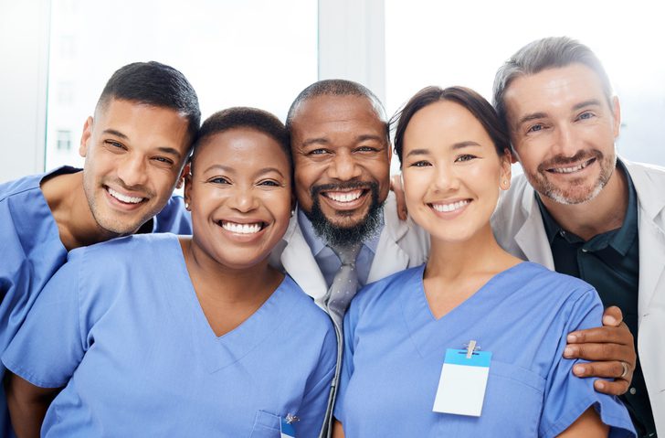 Group of healthcare professionals standing close together in a bright clinical setting, wearing blue medical scrubs and white lab coats, with visible name badges.