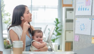 Woman holding a baby while working at a desk in an office, wearing a headset and speaking on the phone, with charts and sticky notes on the wall behind.