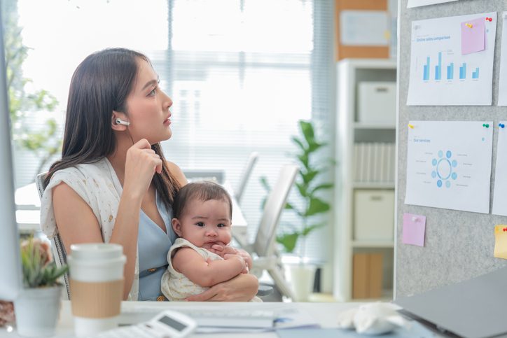 Woman holding a baby while working at a desk in an office, wearing a headset and speaking on the phone, with charts and sticky notes on the wall behind.