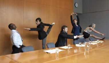 People in business attire doing yoga stretches during a meeting in a conference room.