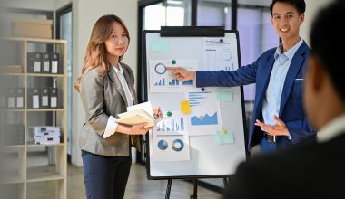Two professionals in an office presenting charts and graphs on a whiteboard, discussing data analysis and project updates in a business setting.