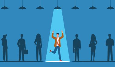 Silhouetted group of six people standing in a row against a blue background. One person in the center is spotlighted with raised arms and lifted leg, appearing celebratory. Only the light above this person is turned on; others remain in shadow.