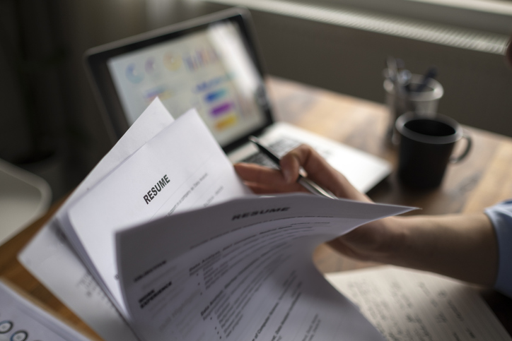 Person holding a resume and a pen in front of a laptop with a coffee mug on the desk