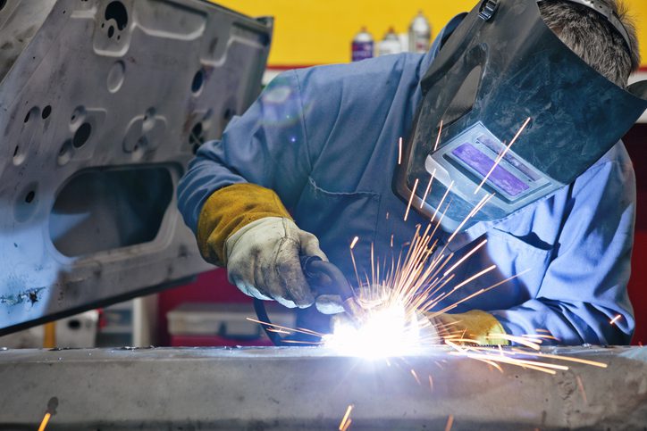 Person welding with protective gear in a workshop, emitting bright light and sparks.