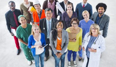 A diverse group of people in professional attire representing occupations like healthcare, construction, engineering, and business, standing in rows and looking up at the camera.