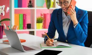 Person in a blue blazer talking on a phone and writing at a desk with a laptop and tablet.