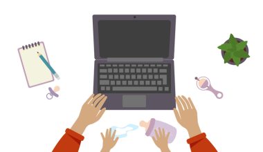 Top-down view of a workspace with a laptop, baby items, and adult and child hands reaching for a bottle.