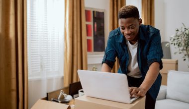 Person working on a laptop placed on cardboard boxes in a room with golden curtains, surrounded by furniture, plants, and wall art.