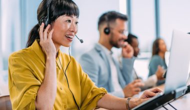 Customer service team in modern office using headsets and laptops for support calls, with natural light from large windows enhancing productivity.