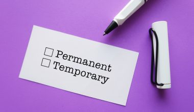 A white card with two checkboxes labeled "Permanent" and "Temporary" is placed on a purple surface. Next to the card, there is a white pen with a black tip and a white cap attached to it by a black string.