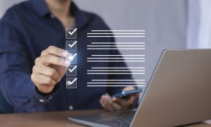 Person in a dark blue shirt using a laptop and holding a smartphone, interacting with a virtual checklist featuring five items marked with checkmarks.