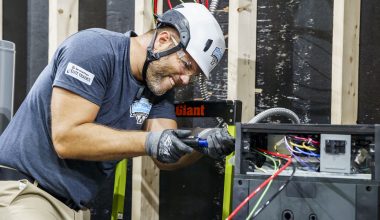 Electrician wearing helmet and gloves working on wiring inside an electrical panel, with visible colored wires.