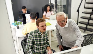 Multigenerational team working in a modern office. An older employee assists a younger colleague at a computer, promoting collaboration and mentorship.