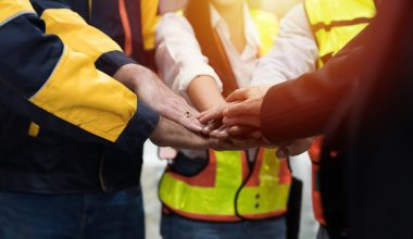 Group of workers in high-visibility safety vests and uniforms standing in a circle with hands stacked together, symbolizing teamwork, unity, and collaboration in an industrial or construction setting. The image reinforces themes of workplace solidarity, team performance, and leadership in hands-on environments.