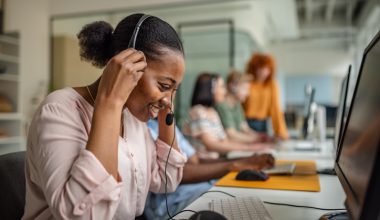 Black woman wearing a headset and working at a computer in a modern office setting, with other team members collaborating in the background.